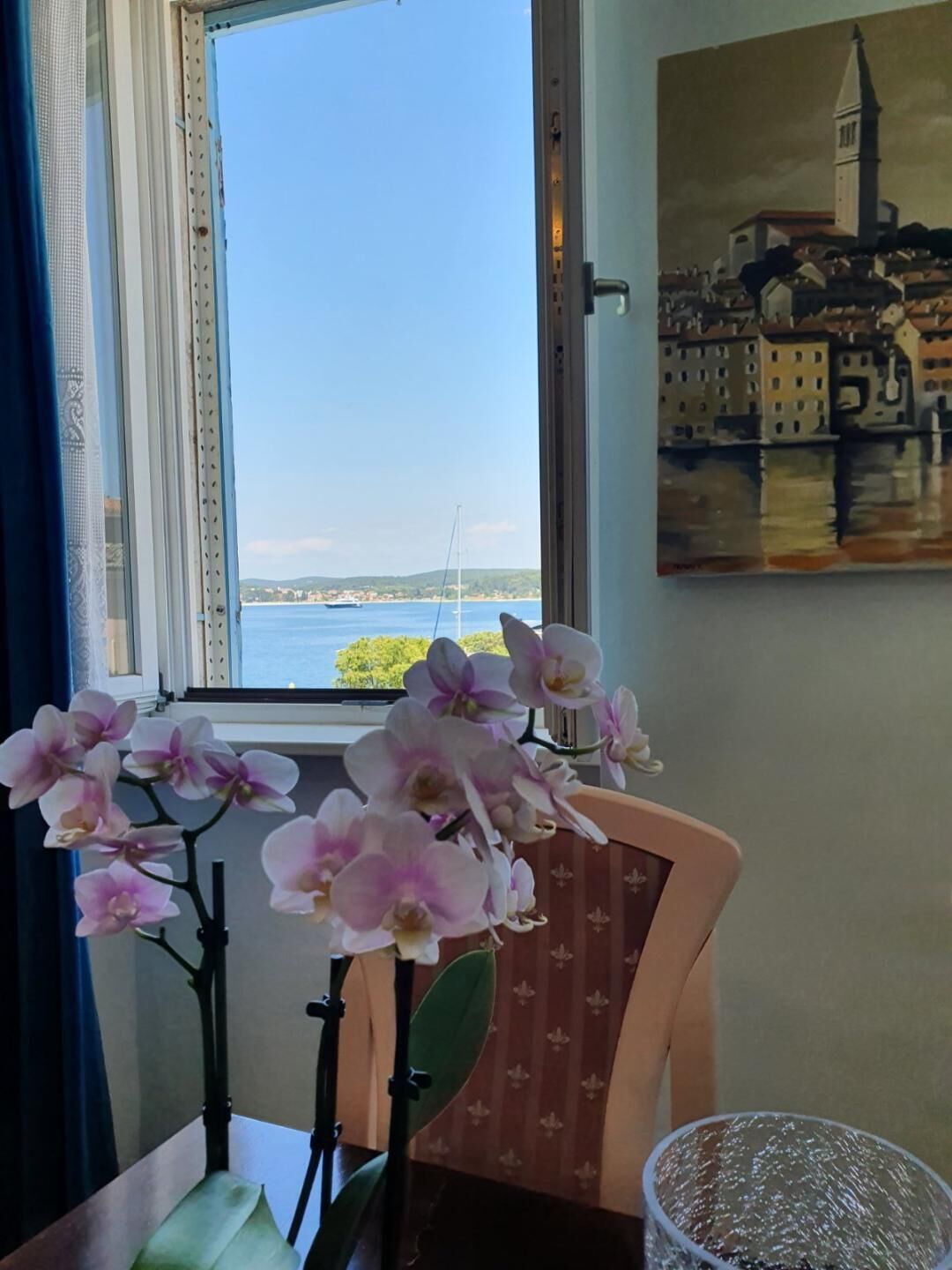 A room with a window overlooking a body of water, a chair with a patterned cushion, and a vase with orchids.
