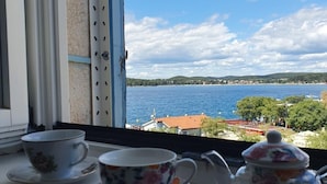 Ett fönster med utsikt över en sjö och byggnader, en kopp, en tekanna och en sockerskål står på fönsterbrädan.