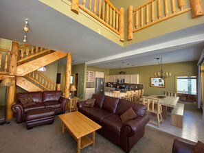 A spacious living area with a wooden staircase, leather sofas, a wooden dining table, and a kitchen in the background.