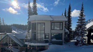Un bâtiment moderne à deux étages, doté de grandes fenêtres et d’un balcon, entouré d’arbres couverts de neige et situé à proximité d’une piste de ski.