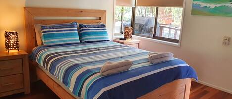 Uma cama com roupa de cama listrada, cabeceira de madeira e duas almofadas. Um criado-mudo com uma luminária e uma gaveta estão presentes. Uma janela com persianas é visível e um quadro está pendurado na parede.