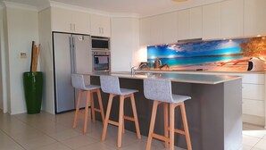 A modern kitchen with a large island, bar stools, a refrigerator, an oven, and a decorative wall mural. - Waterside, Pet Friendly Beach House (Vincentia)