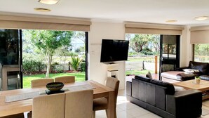 A modern living room with a dining area, a flat-screen TV mounted on the wall, and large sliding glass doors leading to a garden. - Waterside, Pet Friendly Beach House (Vincentia)