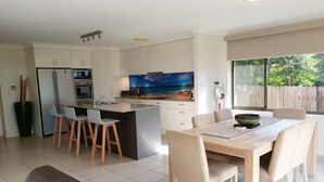 A modern kitchen with a central island, bar stools, and a dining area with a table and chairs. - Waterside, Pet Friendly Beach House (Vincentia)