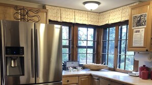Une cuisine avec des meubles de rangement en bois, un réfrigérateur en acier inoxydable, une fenêtre avec des rideaux et une table à manger avec une chaise.