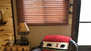 Un fauteuil en cuir avec une couverture, une commode en bois, une lampe et une fenêtre avec des stores.