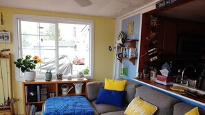 A cozy living room with a grey sofa, blue and yellow cushions, a coffee table, and a bookshelf. A window offers a view of the street.