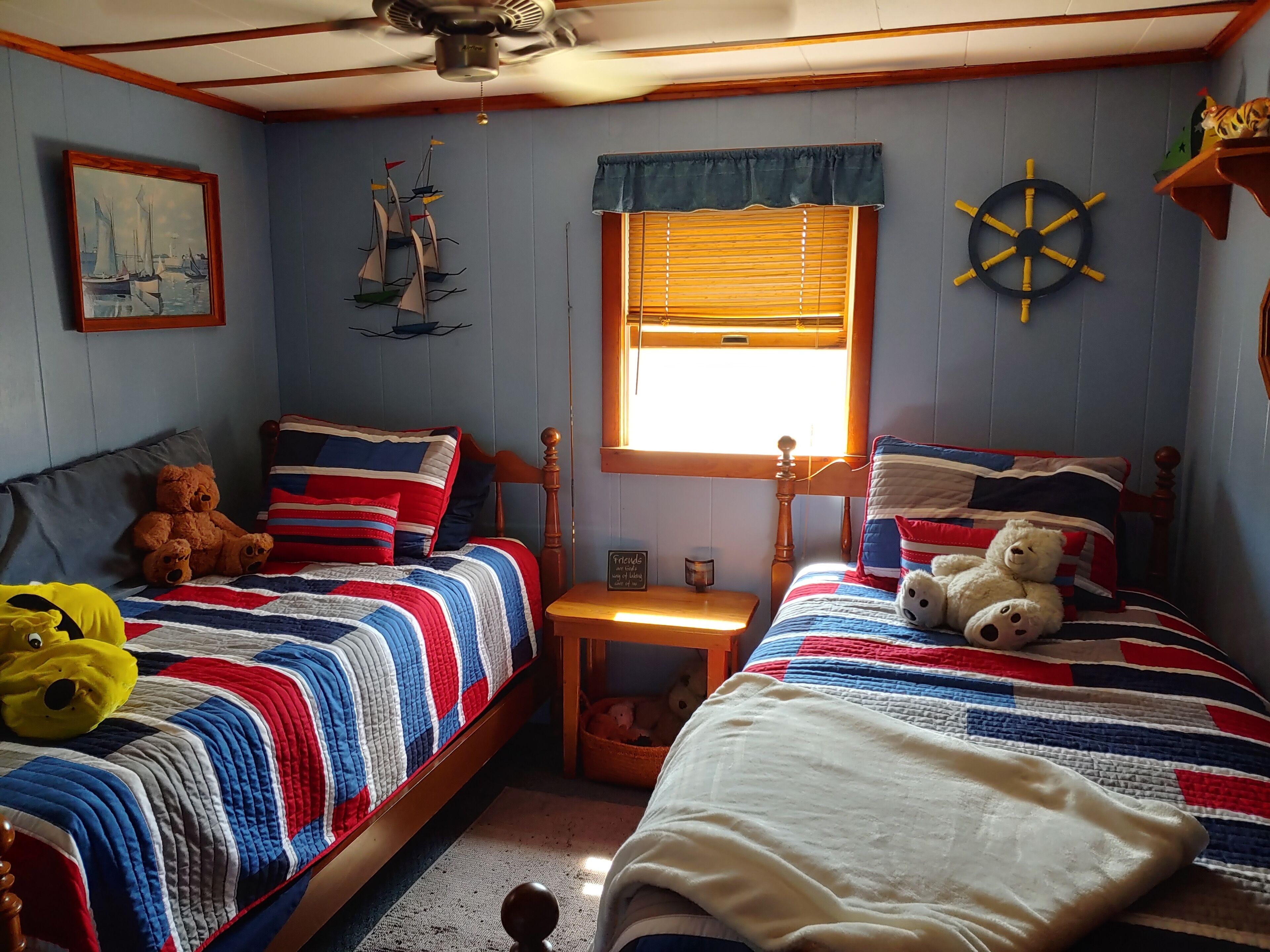 Une chambre avec deux lits, une fenêtre avec des stores, un ventilateur de plafond, une décoration en forme de roue de navire et un ours en peluche sur l’un des lits.