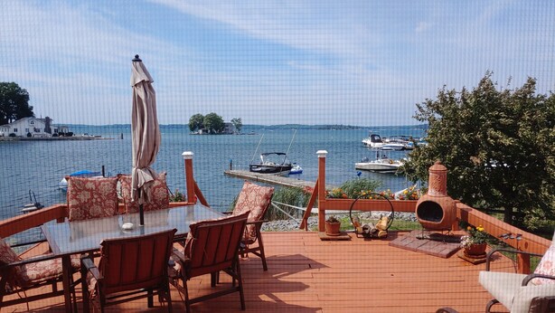 A wooden deck with a table and chairs, overlooking a lake with boats and a house in the distance.