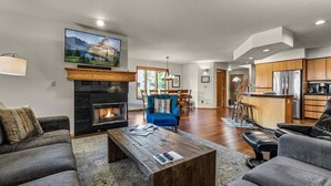 A modern living room with a fireplace, a television mounted above it, and a kitchen area in the background. - Welcome to The Explorers Den, Mountain Harbor #147 (Whitefish)