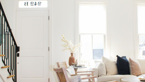A living room with a wooden floor, a white door, a staircase, a sofa, a coffee table, and a vase with flowers.