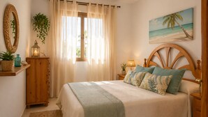 Una camera da letto con un letto, un comodino, una lampada, uno specchio, una finestra con tende e una pianta, oltre a un quadro a tema spiaggia.