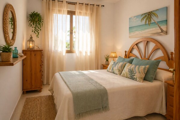 A bedroom with a bed, a nightstand, a lamp, a mirror, a window with curtains, a plant, and a beach-themed painting.