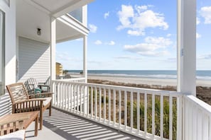 A covered porch with a view of the beach, wooden furniture, and a white railing.