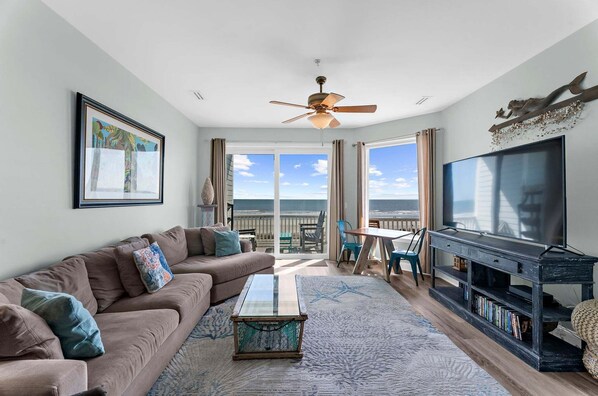 A living room with a sofa, coffee table, TV stand, and a view of the ocean through large windows.