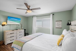 Un dormitorio con cama, ventilador de techo, televisor, cómoda, ventana con persianas y una frase motivadora en la pared.