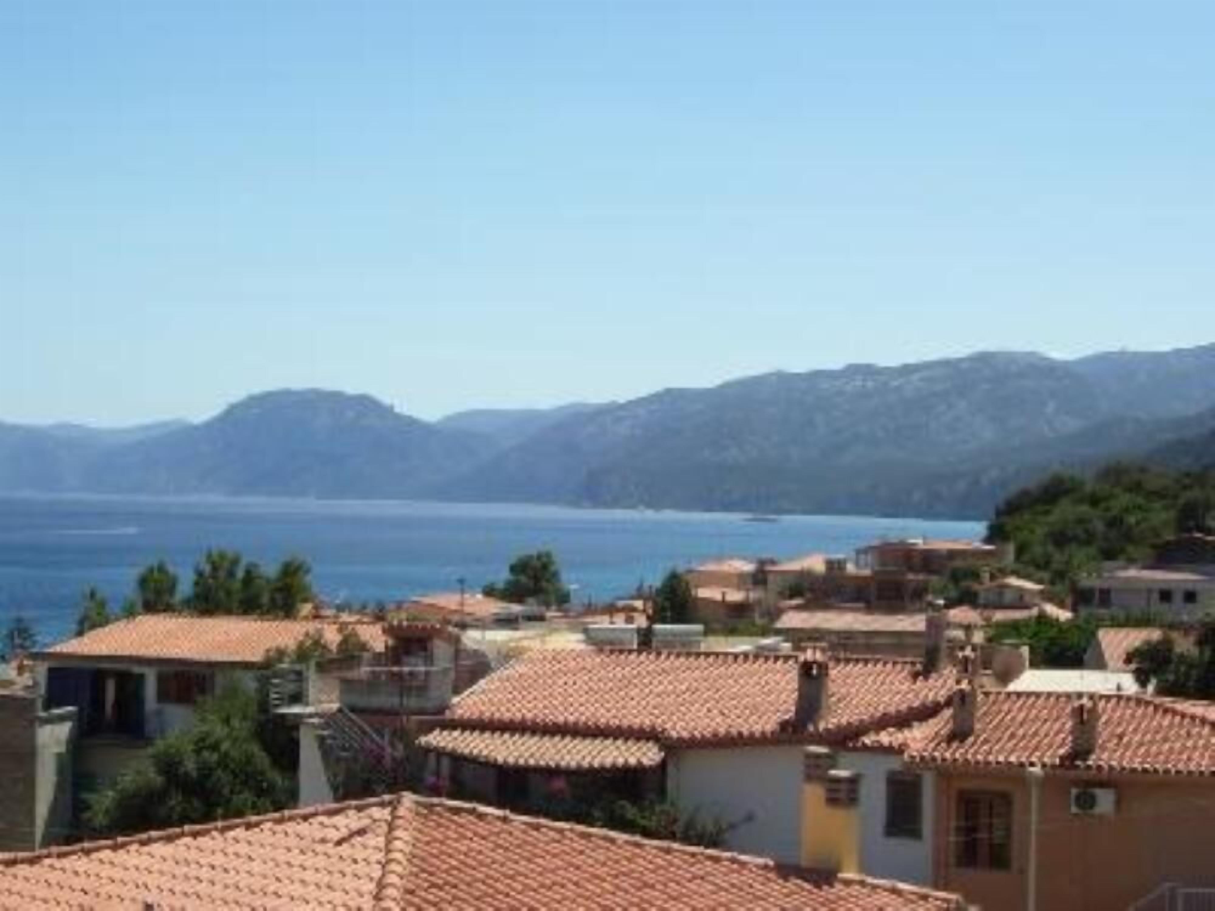 A coastal village with terracotta-roofed buildings, a clear view of the sea, and mountains in the background.