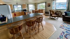 A spacious kitchen with a central island, wooden bar stools, and a dining table with chairs. There is a large window providing a view of the outside.