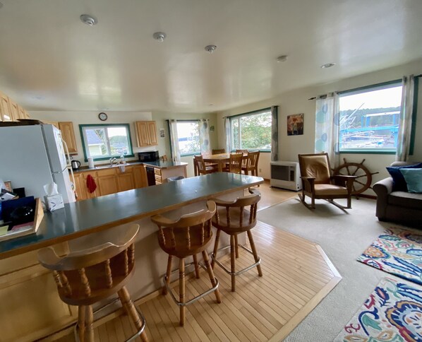 A spacious kitchen with a central island, wooden bar stools, and a dining table with chairs. There is a large window providing a view of the outside.