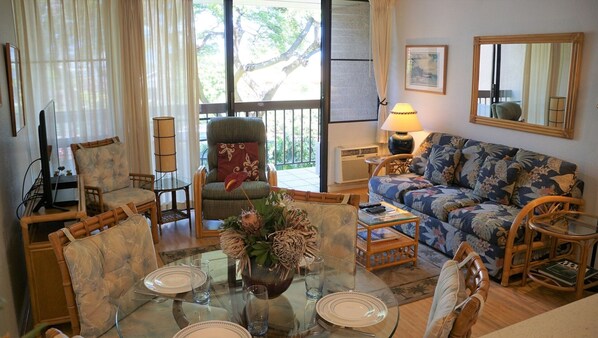 A living room with a glass dining table set for four, surrounded by wicker furniture, a sofa with a floral pattern, and a chair with a red cushion.