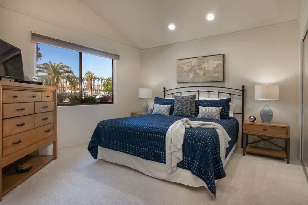 Een slaapkamer met een groot bed, een houten dressoir, een nachtkastje en een raam met uitzicht op palmbomen.