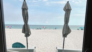 Vista a la playa desde una habitación, con una silla blanca y una mesa, un paraguas estampado, y la playa visible con gente y paraguas.