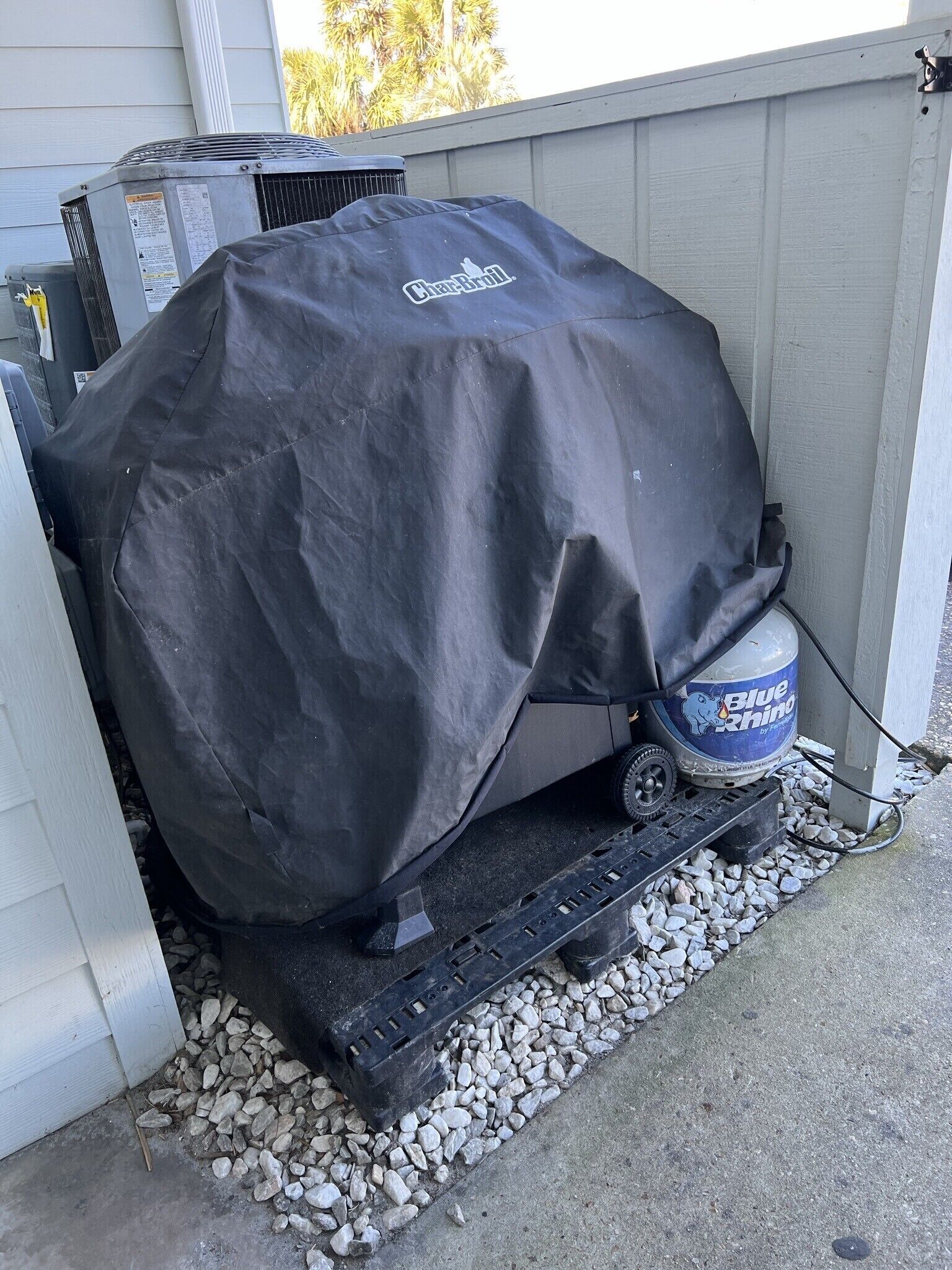 Een barbecue afgedekt met een zwarte beschermkap, een propaantank en een grillmat op een betonvloer met grind.