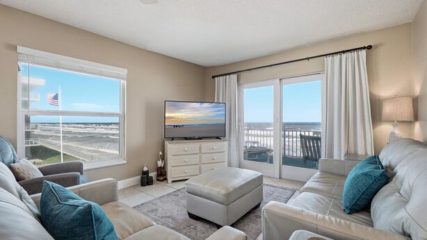 A living room with a television, a sofa, an ottoman, and a view of the ocean. - Bright oceanfront corner 2/2 with beach/pool views, large balcony, and multiple pools (New Smyrna Beach)