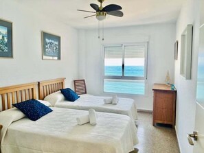 Een kamer met twee bedden, een plafondventilator, een raam met uitzicht op zee en een klein houten kastje.