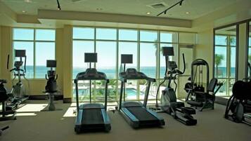 A gym with treadmills, elliptical machines, and exercise bikes, featuring large windows with a view of the ocean.