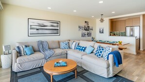 A modern living room with a sectional sofa, a coffee table, and a kitchen area in the background. - Seahorse Retreat - Double The Fun, BEACHFRONT, In Carpinteria! (Carpinteria)