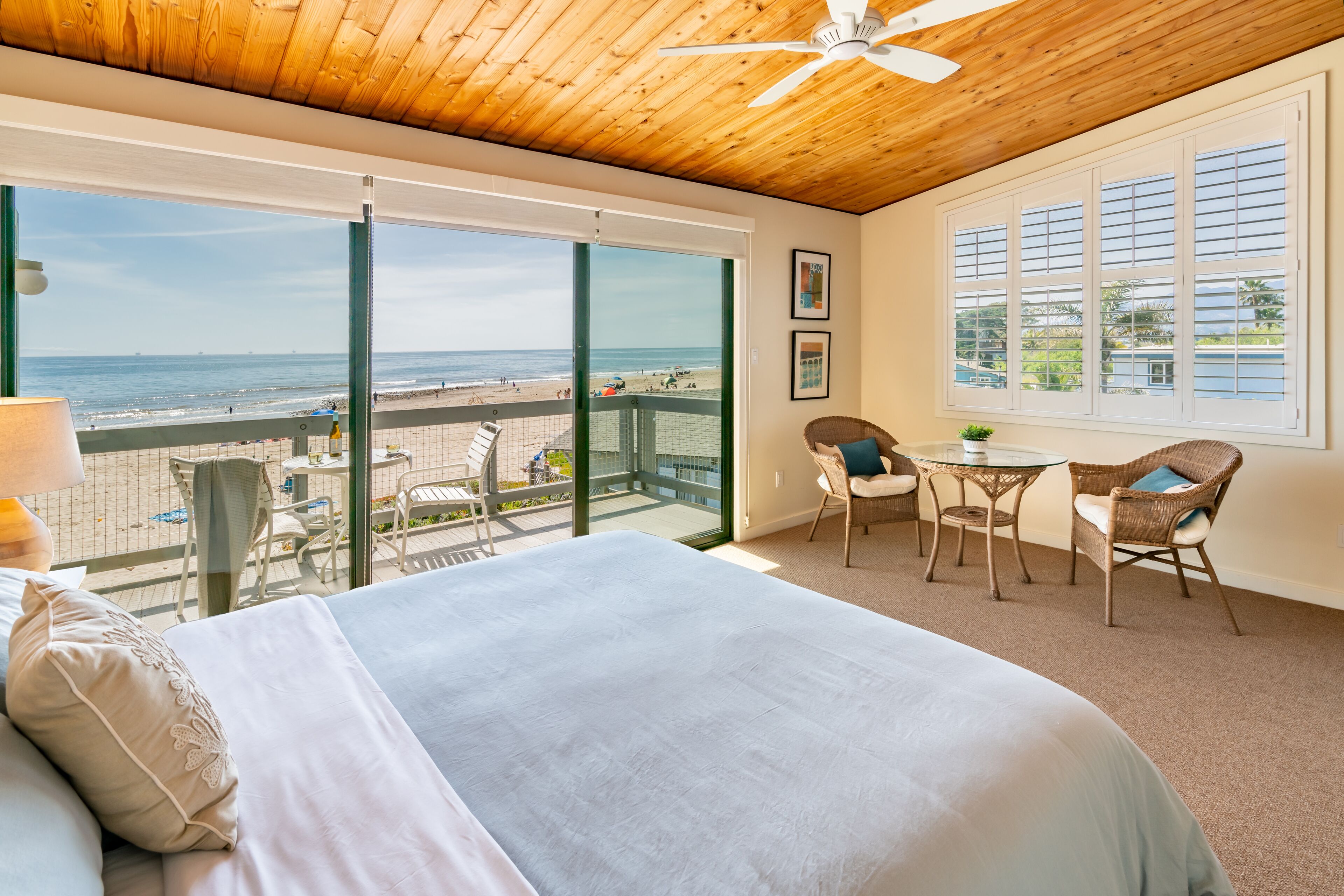 Un dormitorio con una cama grande, un balcón con vista a la playa, sillas de mimbre y un ventilador de techo.