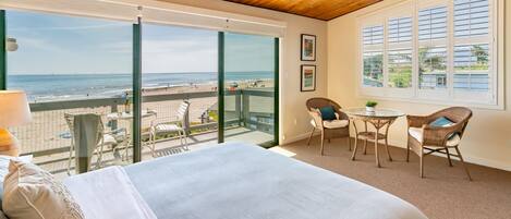 Un dormitorio con una cama grande, un balcón con vista a la playa, sillas de mimbre y un ventilador de techo.