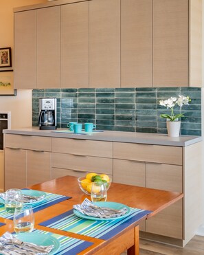 A modern kitchen with a coffee maker, a bowl of fruit, and a dining table set with plates and glasses. - Seahorse Retreat - Double The Fun, BEACHFRONT, In Carpinteria! (Carpinteria)