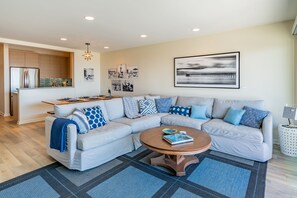 A modern living room with a sectional sofa, a coffee table, and a kitchen area in the background. - Seahorse Retreat - Double The Fun, BEACHFRONT, In Carpinteria! (Carpinteria)