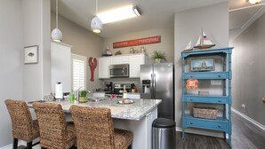 A kitchen with a central island, white cabinets, stainless steel appliances, wicker chairs, and a blue shelving unit. - Read Our Reviews Gorgeous 2 BR Poolside T/h, 3 minute walk to beach! (Corpus Christi)