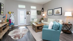 A living room with a teal armchair, a white sofa, a wooden coffee table, and a small side table with a lamp. There is a door with a window, a wall-mounted shelf with decorative items, and a rug on the floor. - Read Our Reviews Gorgeous 2 BR Poolside T/h, 3 minute walk to beach! (Corpus Christi)