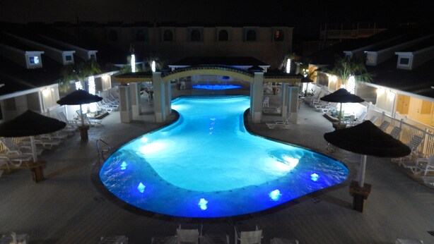 A nighttime swimming pool with a curved design, surrounded by lounge chairs and umbrellas, and a covered seating area in the background. - Read Our Reviews Gorgeous 2 BR Poolside T/h, 3 minute walk to beach! (Corpus Christi)