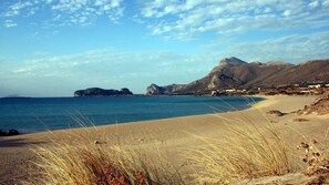 Ein Strand mit klarem, blauem Wasser, sandigem Ufer und hügeliger Kulisse im Hintergrund.