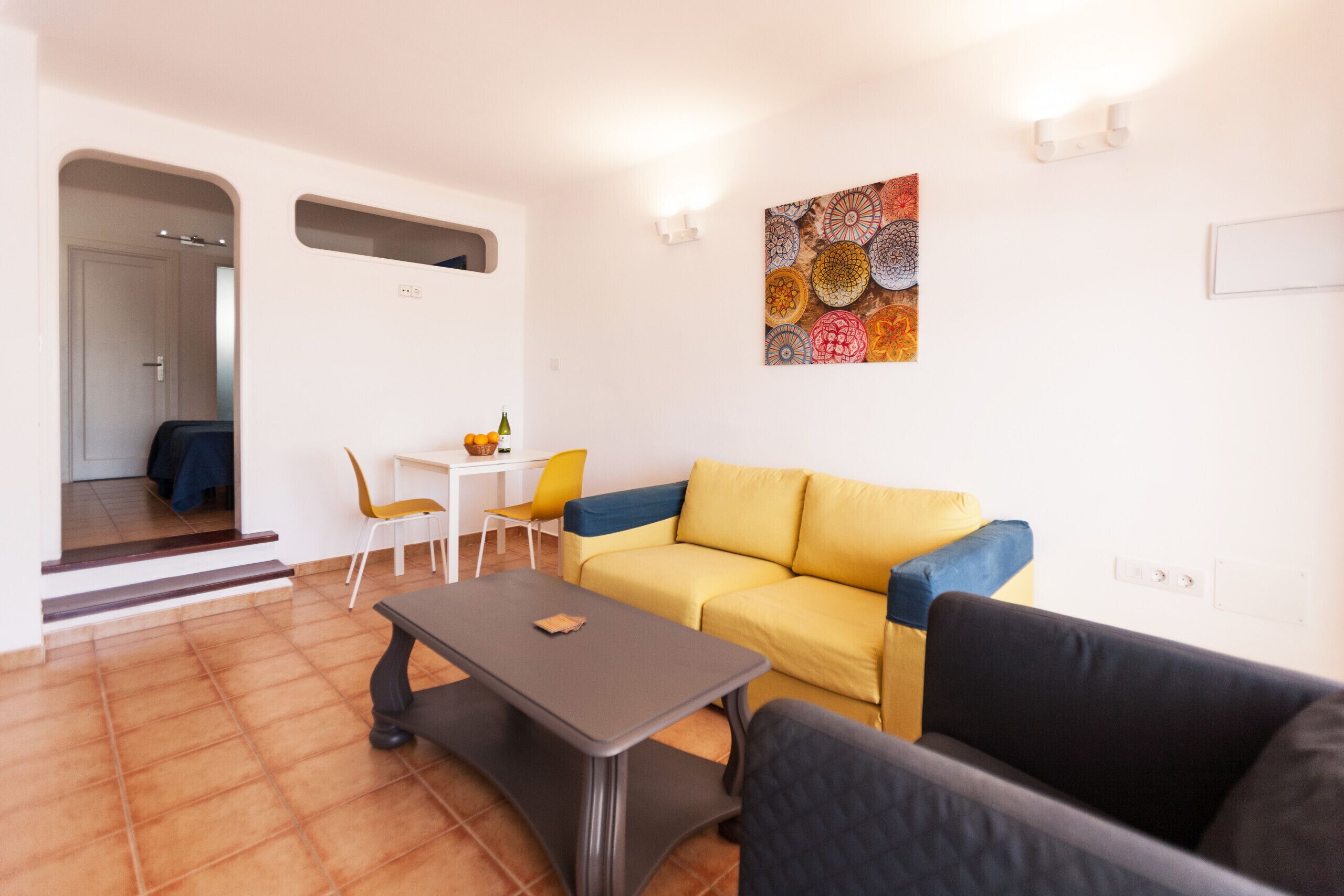 Un salon avec un canapé jaune, un fauteuil sombre, une petite table et un mur blanc orné d’une œuvre d’art colorée.