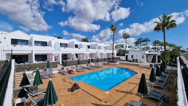 A pool area with lounge chairs and umbrellas, surrounded by white buildings with balconies and palm trees. - Princesa Ico 1 bedroom apts. (Puerto del Carmen)