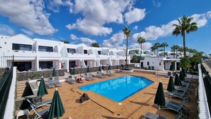 A pool area with lounge chairs and umbrellas, surrounded by white buildings with balconies and palm trees. - Princesa Ico 1 bedroom apts. (Puerto del Carmen)