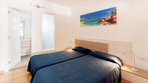 A bedroom with two beds, a wooden headboard, a nightstand with a lamp, and a painting of a beach scene on the wall. - Princesa Ico 1 bedroom apts. (Puerto del Carmen)