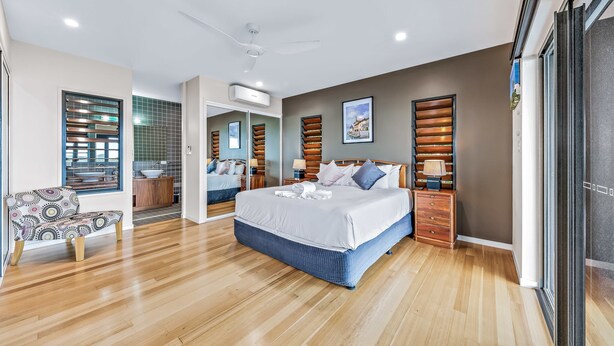 A modern bedroom with a large bed, a wooden bench, and a view of the bathroom. - Shutehaven Villa (Shute Harbour)