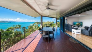 A spacious balcony with a wooden floor, a dining table with chairs, a ceiling fan, and a TV in the background. - Shutehaven Villa (Shute Harbour)