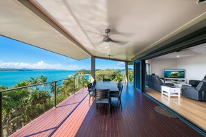 A spacious balcony with a wooden floor, a dining table with chairs, a ceiling fan, and a TV in the background.