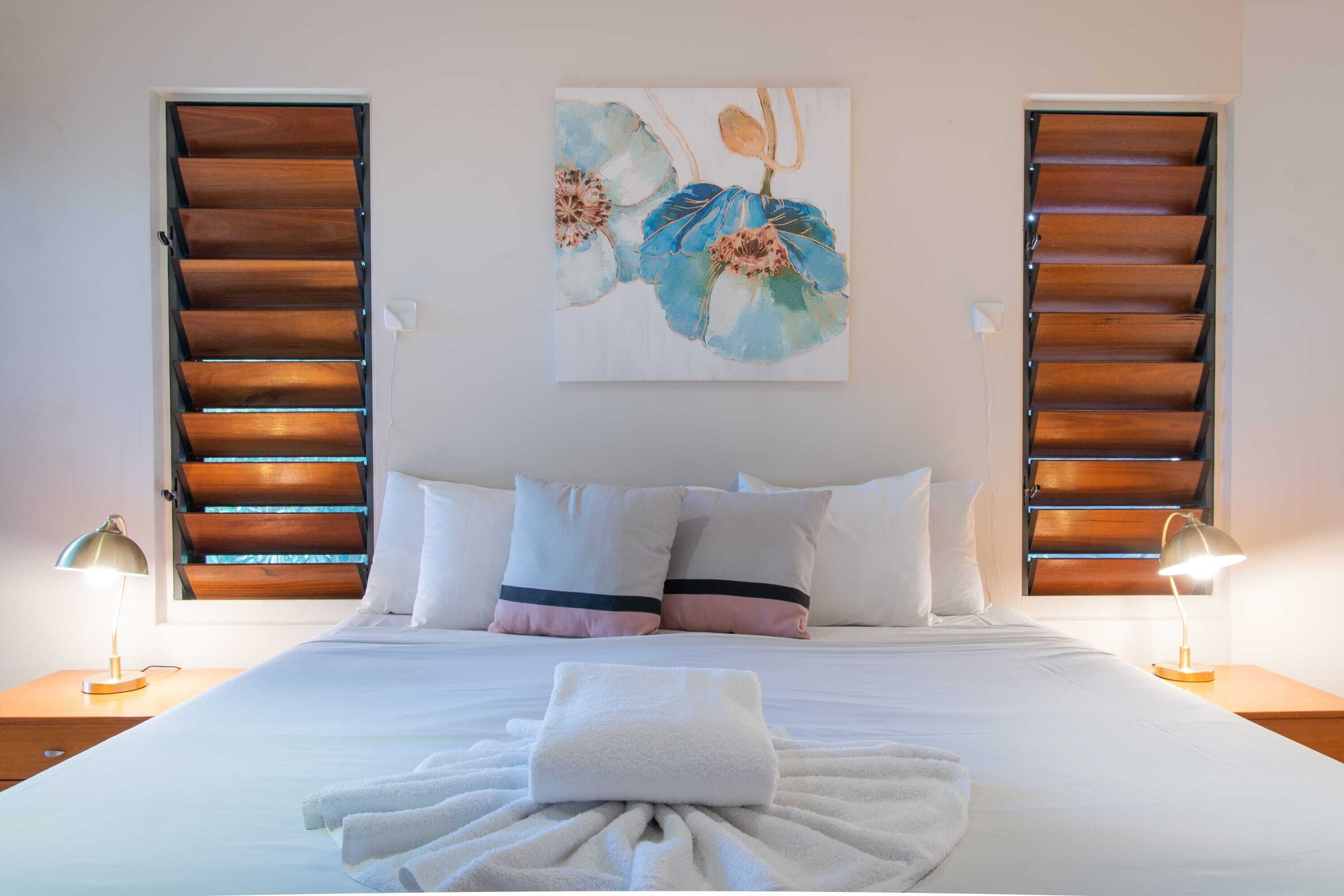 A neatly made bed with white linens, two bedside lamps, and a painting of flowers on the wall.