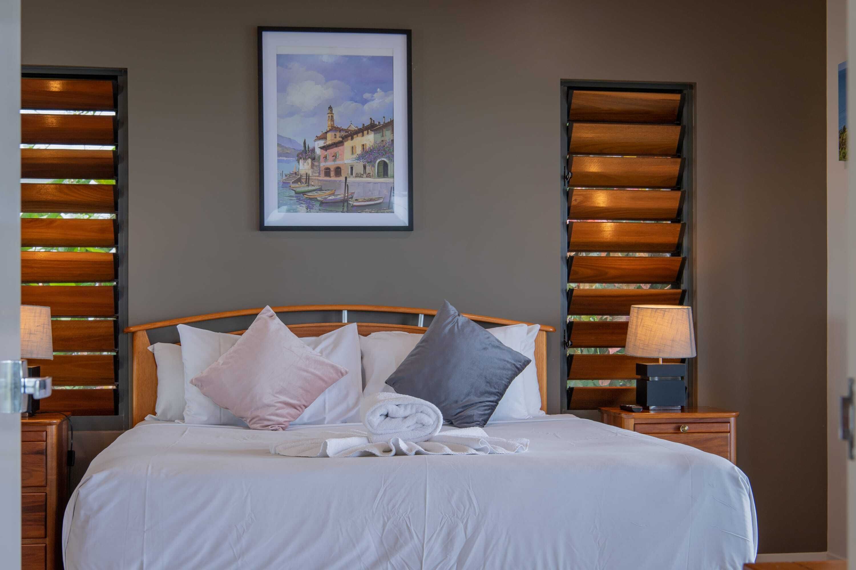 Una cama bien hecha con almohadas, un cuadro en la pared y persianas de madera.