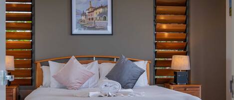A neatly made bed with pillows, a framed picture on the wall, and wooden slat blinds on the windows.