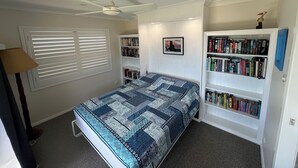 Een slaapkamer met een bed, een plafondventilator, een boekenplank en een raam met jaloezieën.